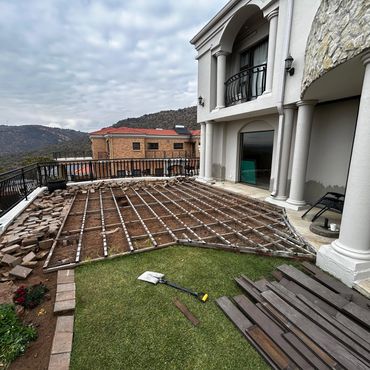 Patio renovation with removed tiles and visible wooden frame beside a house.