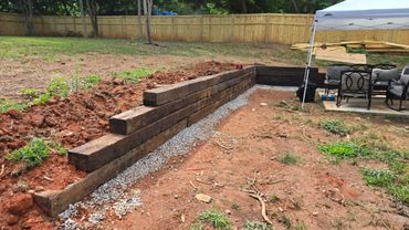 Construction of a wooden retaining wall in a backyard with gravel base and patio furniture nearby.