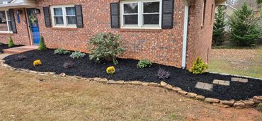 Freshly mulched garden bed with assorted shrubs and stone edging along a brick house.