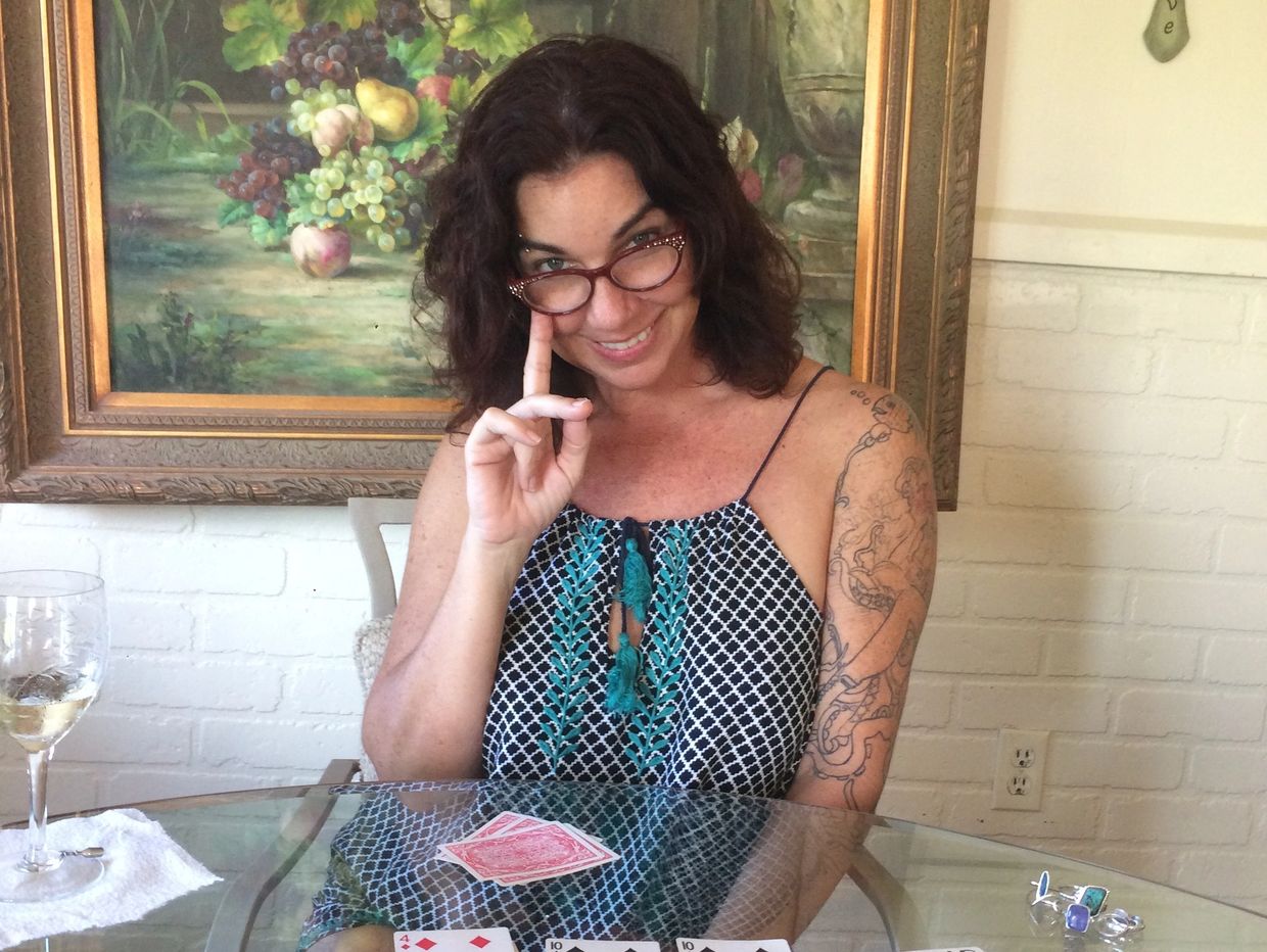 Woman with glasses sitting at a glass table with playing cards, smiling warmly.