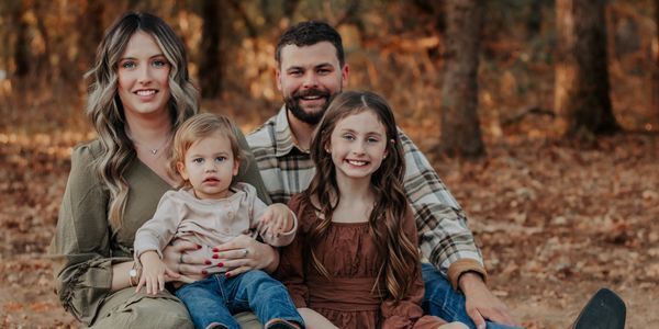 A family of four posing outdoors on autumn leaves, smiling warmly.