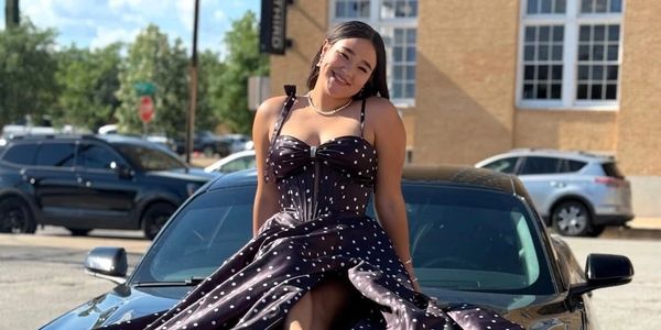 A smiling woman in a black dress with white dots sits on a car hood on a sunny day.