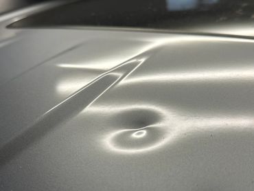 Close-up of a dent on a silver car hood under bright lighting.