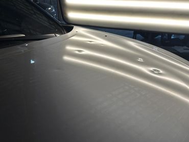 Car hood with multiple dents and reflections from overhead lights.
