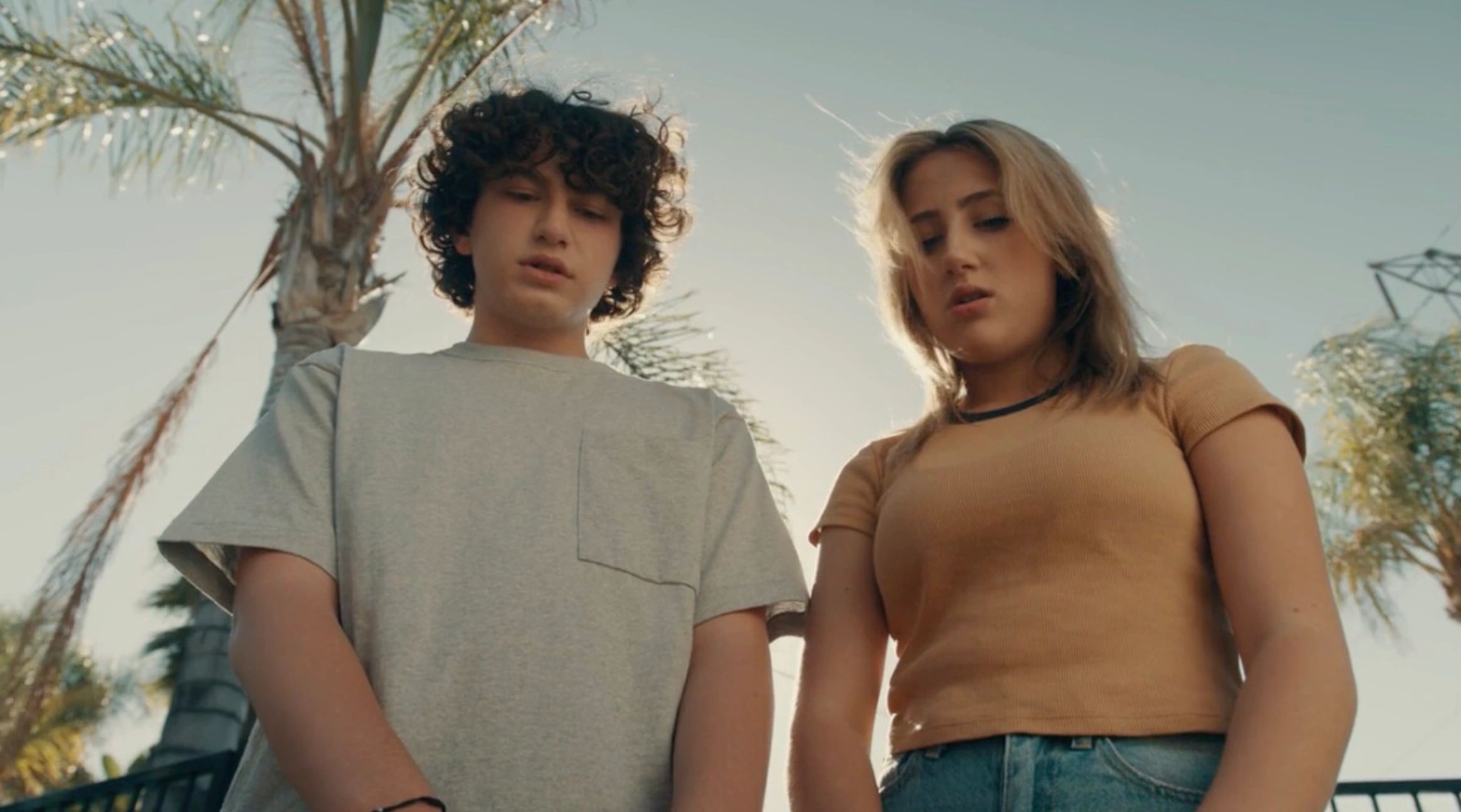 Two young people looking down with concerned expressions outdoors under palm trees.