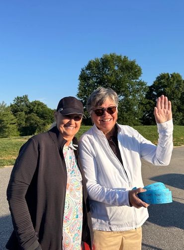 Two smiling women outdoors on a sunny day, one holding tickets and waving.