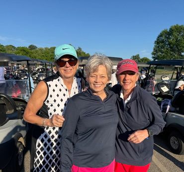 Three women smiling at a golf course parking area with golf carts behind them.