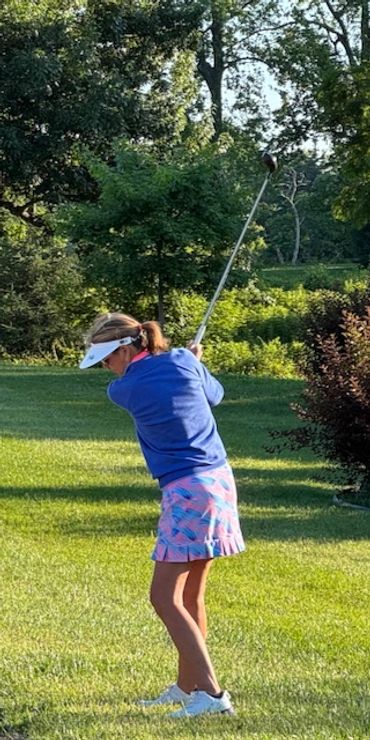 Woman in blue sweater and pink skirt swinging a golf club on a sunny course.