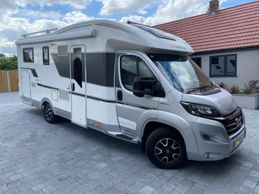 White and gray Adria motorhome parked on a paved driveway beside a house.