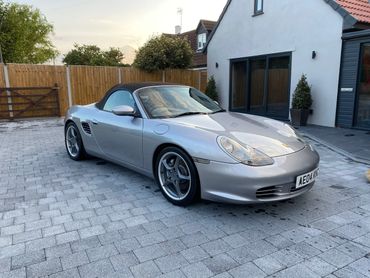 Silver Porsche Boxster convertible parked on a paved driveway beside a modern house.