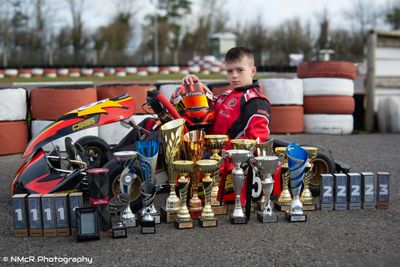 Young go-kart racer poses with trophies and kart on track.