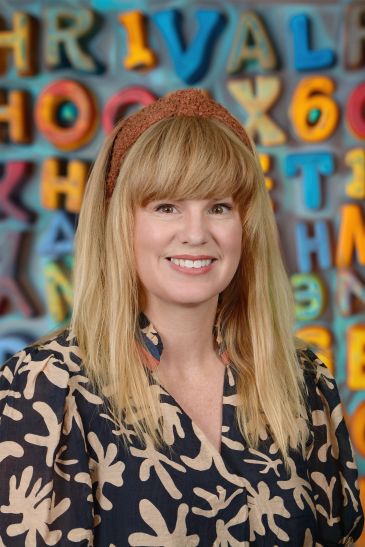 Smiling woman with blonde hair stands in front of colorful alphabet wall.