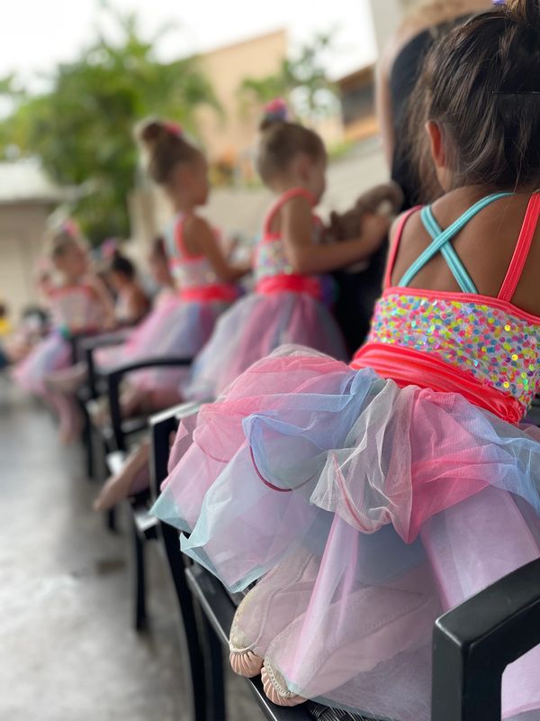 Young ballerinas in colorful tutus sitting in a row on chairs.