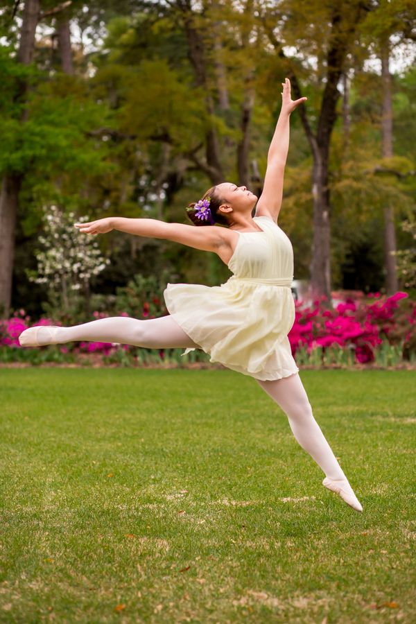 Ballerina in a white dress leaps gracefully outdoors with flowers in her hair.