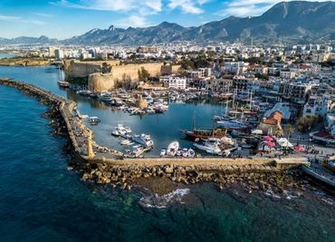 A coastal city harbor with boats and mountainous backdrop under a blue sky.