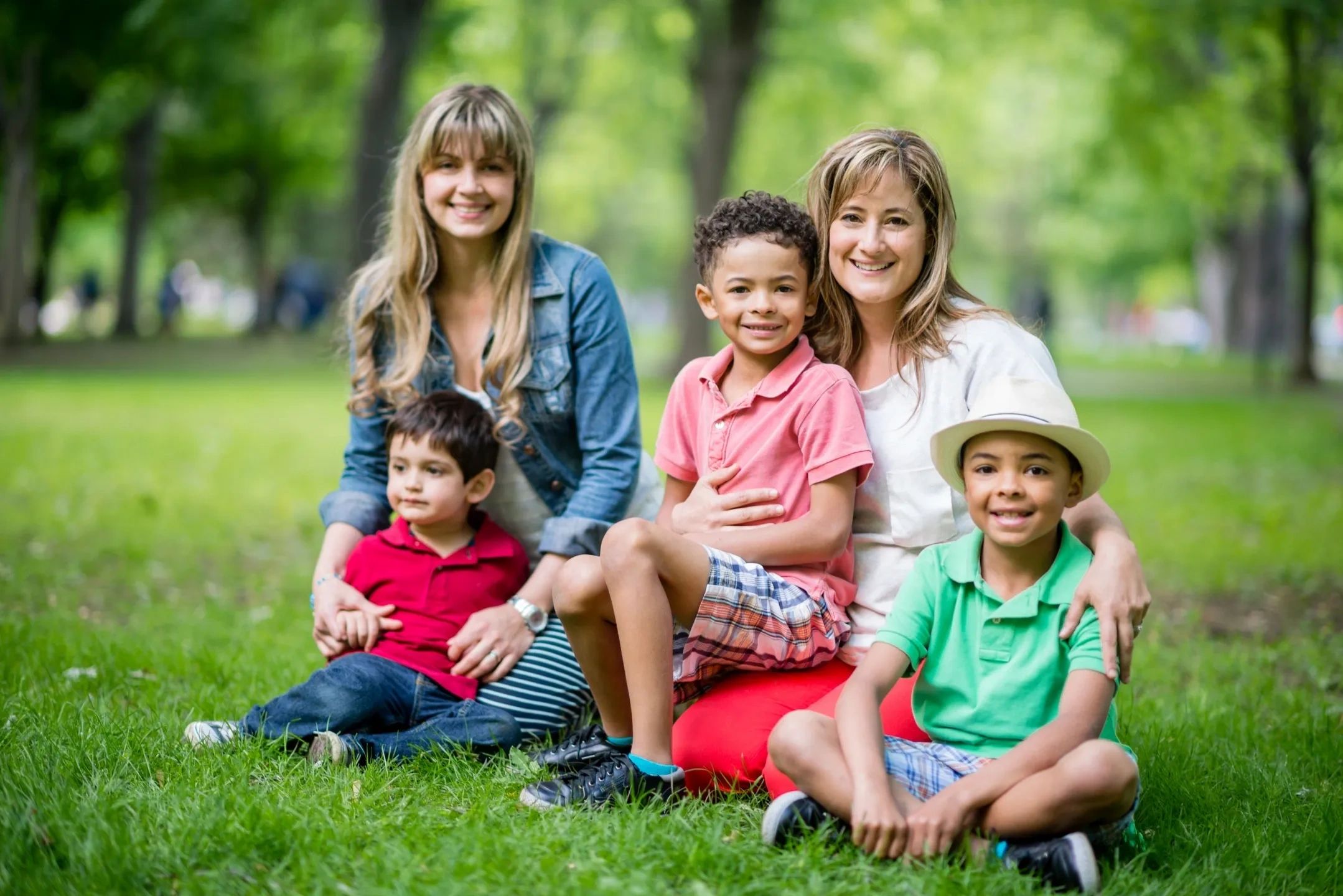 Two women with three kids sitting on grass in a park on a sunny day.