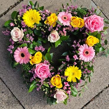 Heart-shaped floral wreath with pink, yellow, and white flowers on pavement.