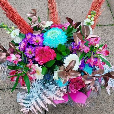 Vibrant floral arrangement with blue, pink, and white flowers accented by copper leaves and orange pampas grass.