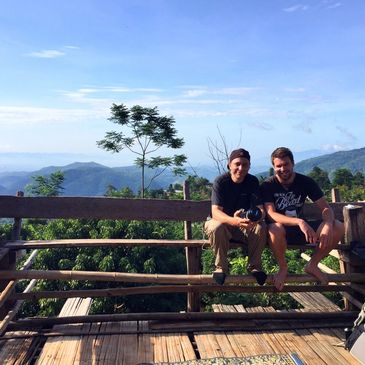 Two men sitting on a wooden bench with mountainous landscape behind them.