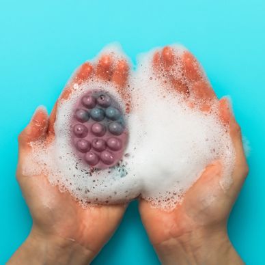 Two open hands holding lather with an oval purple and blue bar soap on the left hand.
