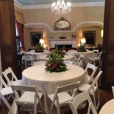Elegant room with round tables, white chairs, and floral centerpieces under a chandelier.