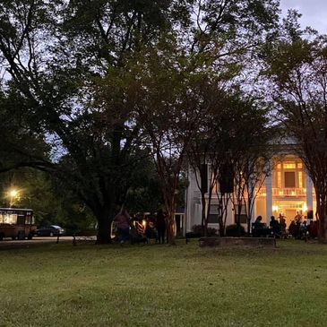 Evening gathering at a lit historic white mansion with trees around.