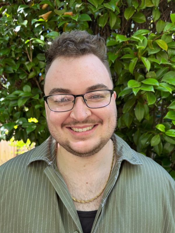 Smiling man with glasses in front of green leafy background.
