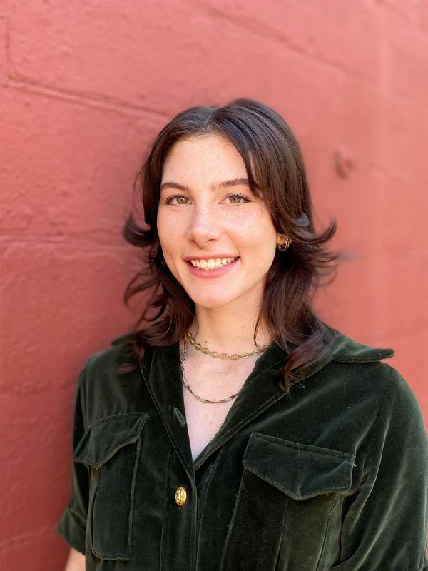 A young woman with brown hair smiles against a red brick wall.