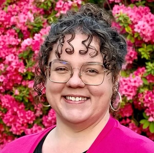 Smiling woman with glasses and curly hair in front of pink flowers.