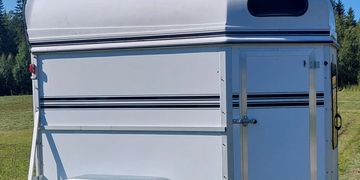 White horse trailer parked on grass with trees in the background.