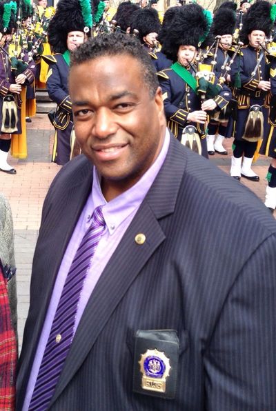 Man in suit with badge at parade with bagpipers in traditional attire.