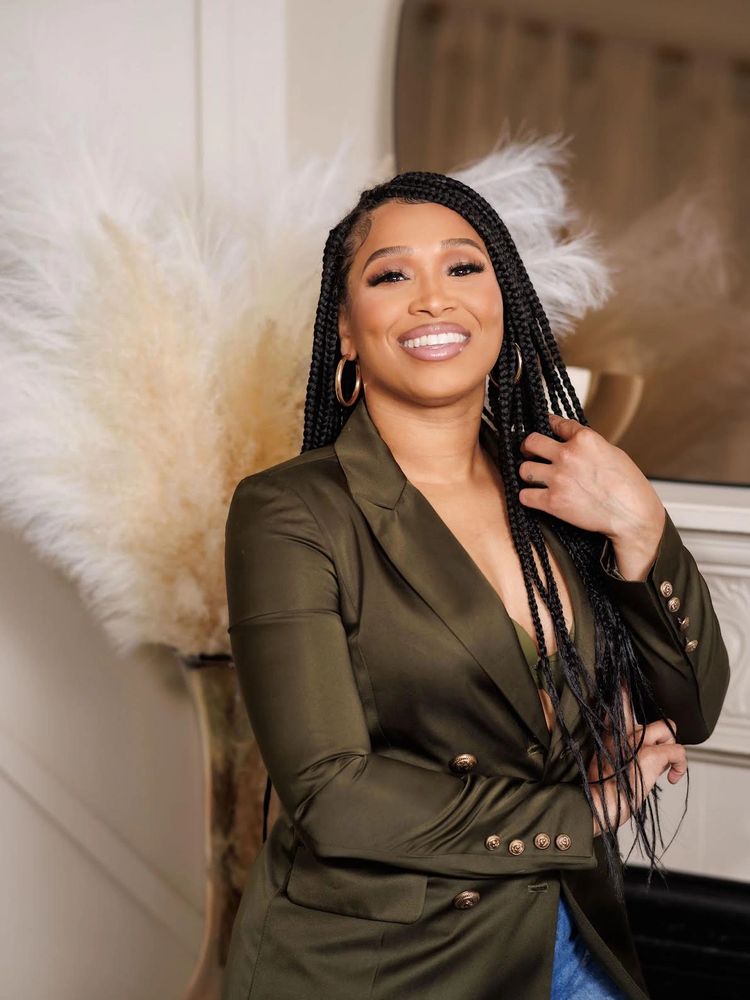 Confident woman with braided hair smiles, wearing an olive blazer and gold hoop earrings.