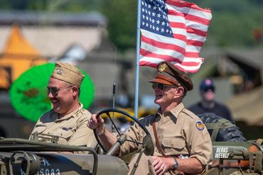 Two men in uniform driving WWII car. American flag is flying in the background.