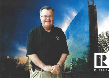 Smiling man in black polo stands outdoors with cityscape and Capitol building at sunset.
