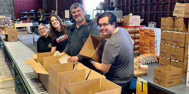 Four people packing boxes in a warehouse with smiles on their faces.