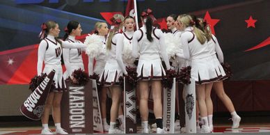 Cheerleaders in white uniforms gather in a huddle with pom-poms and megaphones.