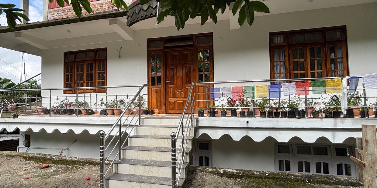 A white house with wooden doors and windows, stairs leading to the porch, and prayer flags hanging on the railing.
