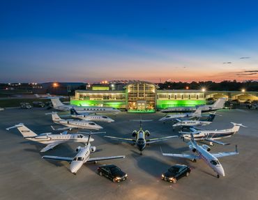 Luxury private jets and cars parked in front of a modern airport terminal at dusk.