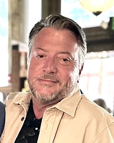 Smiling man with gray hair and beard wearing a beige jacket indoors.