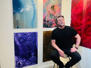 Man sitting on a stool in an art gallery with colorful abstract paintings.