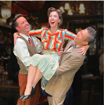 Two men lift a joyful woman dressed in vintage attire in a lively pub.