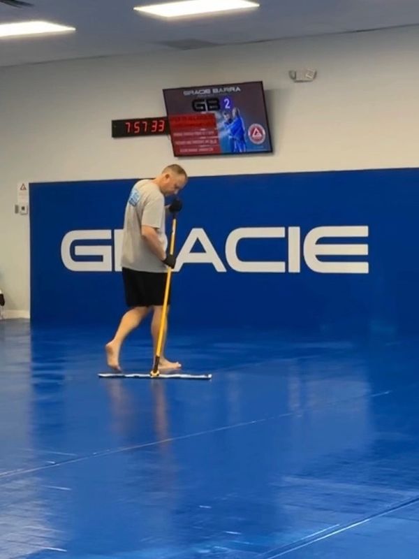 A man cleans a blue mat in a martial arts gym with a mop.