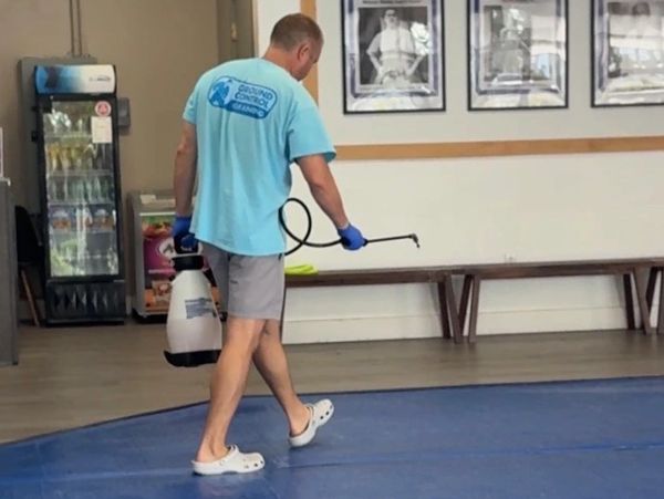 Man spraying disinfectant on a blue mat indoors, wearing casual clothes and gloves.