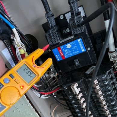 Hand holding a yellow digital clamp meter measuring voltage inside an electrical panel.