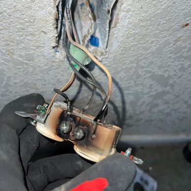 Close-up of a burnt electrical outlet with exposed wires held by a gloved hand.