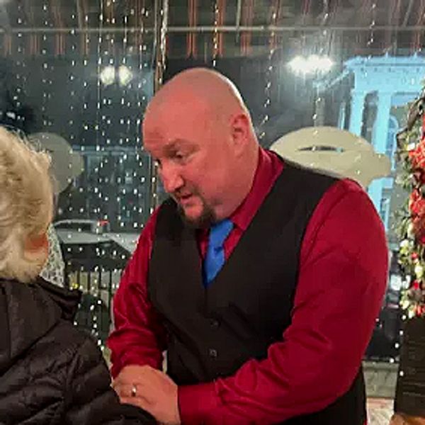 Bald man in a red shirt and black vest talking to a woman indoors.