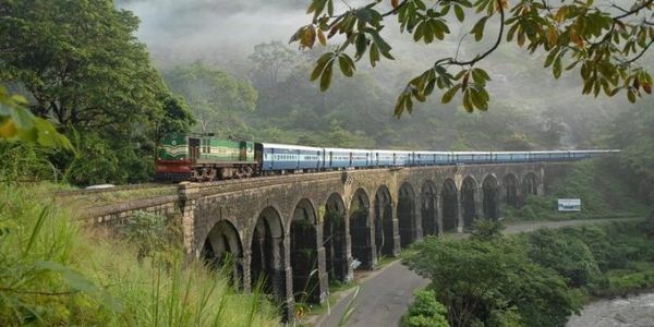 A train crosses a historic arched stone bridge surrounded by lush greenery and misty hills.