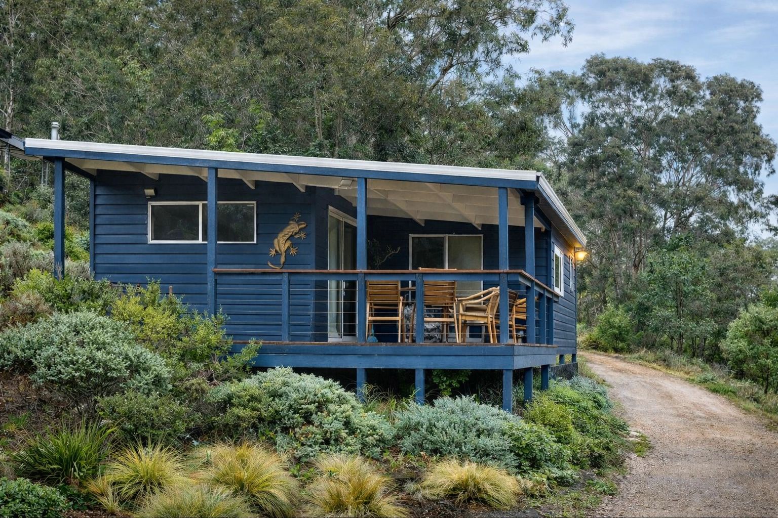 Blue cabin with wooden porch chairs near a dirt path surrounded by greenery.