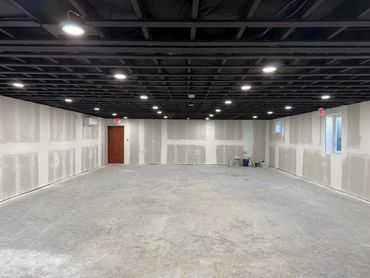 Empty basement room with unfinished drywall and exposed black ceiling beams.