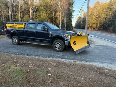 Black pickup truck with a yellow snowplow attached, parked by the roadside.